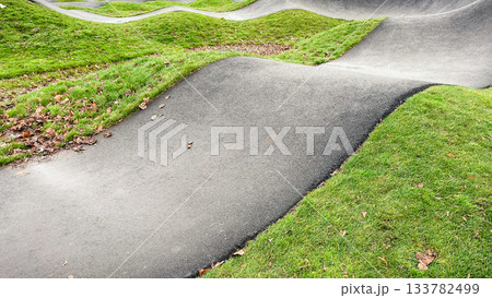 Curved asphalt pump track for cycling and wheeled sports, surrounded by vibrant green grass in a modern park 133782499