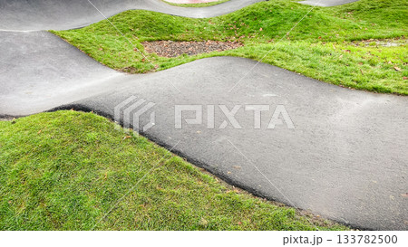 Curved asphalt pump track for cycling and wheeled sports, surrounded by vibrant green grass in a modern park 133782500