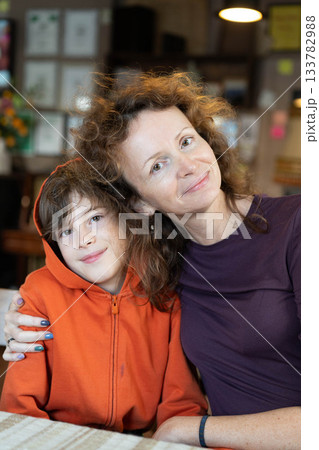 A mother and her son share a warm embrace at a dinner table. They smile joyfully in a relaxed restaurant, surrounded by charming decor, showcasing a heartwarming moment of connection. 133782988