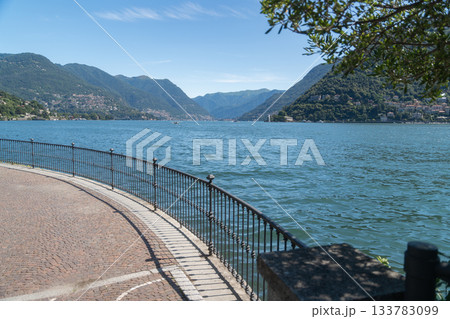 A curved stone promenade lines the lake, with mountains in the background and tree branches providing soft shade. A bright, calm summer scene with a pleasant sense of openness., Italy, Como A curved stone promenade lines the lake, with mountains in the background and tree branches providing soft shade. A bright, calm summer scene with a pleasant sense of openness., Italy, Como 133783099