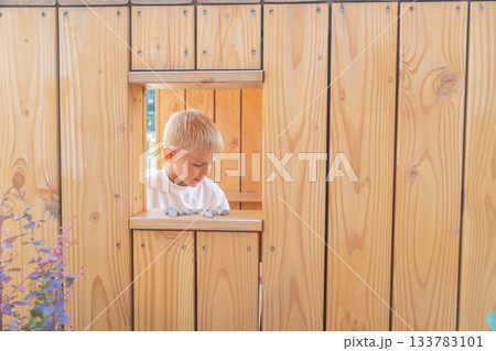A young boy stands inside a wooden play structure, looking through a small window. Warm natural tones and soft daylight create a calm outdoor scene with a simple, rustic and playful atmosphere 133783101
