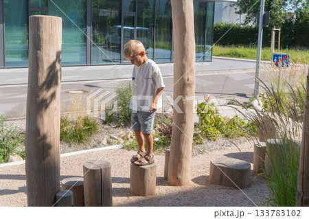 A boy walks along wooden stepping logs in an outdoor natural play area. The scene combines sunlight, rustic materials, greenery, and gentle motion, conveying exploration, balance, and a calm summer A boy walks along wooden stepping logs in an outdoor natural play area. The scene combines sunlight, rustic materials, greenery, and gentle motion, conveying exploration, balance, and a calm summer 133783102