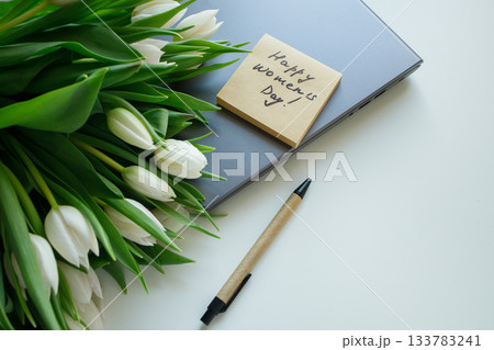 Sticky note with Women's day handwritten congratulations and white tulips bouquet laying on the laptop at white office table. Greetings for women at workplace. Sticky note with Women's day handwritten congratulations and white tulips bouquet laying on the laptop at white office table. Greetings for women at workplace. 133783241