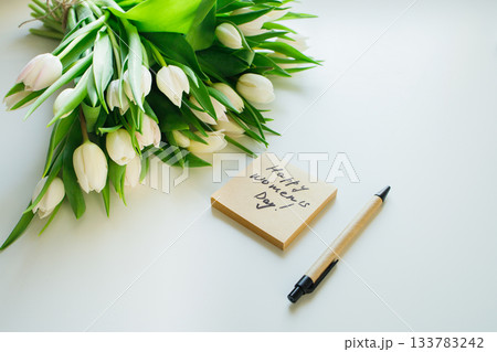 Sticky note with Women's day handwritten congratulations and white tulips bouquet laying at white office table. Greetings for women at workplace. Sticky note with Women's day handwritten congratulations and white tulips bouquet laying at white office table. Greetings for women at workplace. 133783242