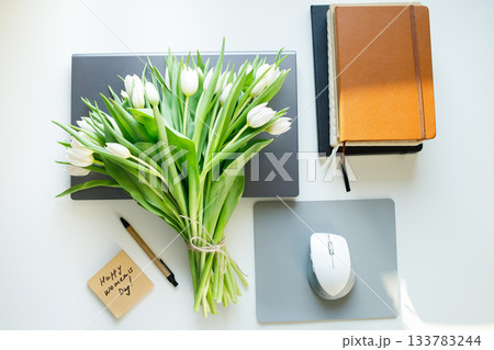 Sticky note with Women's day congratulations and white tulips bouquet laying at office workplace table with laptop, computer mouse, notepads. Sunny spring holiday morning greetings from colleagues. 133783244