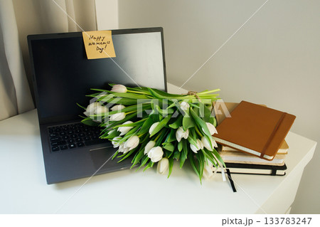 Surprise sticky note with Women's day handwritten congratulations left on the laptop and fresh white tulips bouquet lying at woman workplace. 133783247