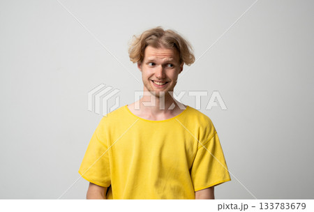 Smiling young man looking sideways with awkward amused expression while posing in neutral studio scene Smiling young man looking sideways with awkward amused expression while posing in neutral studio scene 133783679