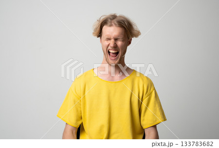 Young man in yellow shirt screaming loudly with eyes closed and emotional expression in studio portrait 133783682