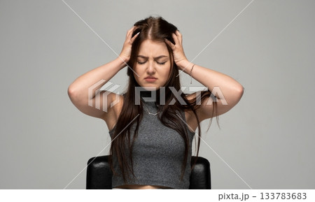 Stressed young woman holding her head with both hands and closing eyes while sitting in studio scene Stressed young woman holding her head with both hands and closing eyes while sitting in studio scene 133783683