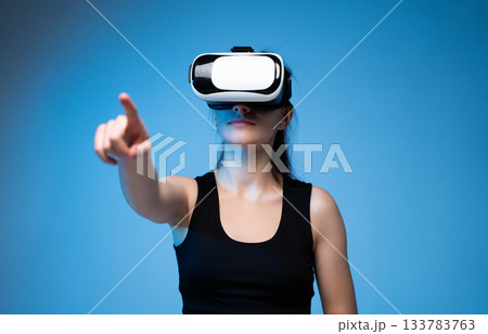 Woman wearing VR headset and interacting with virtual environment while pointing forward in blue studio 133783763