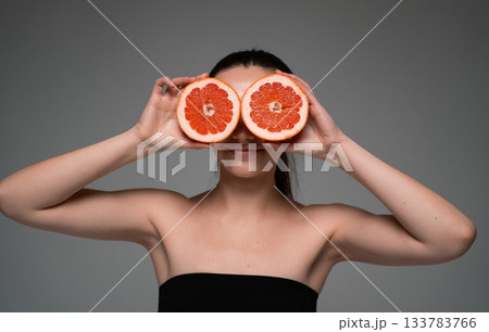 Cheerful woman holding two grapefruit slices over eyes, representing citrus freshness, fun energy and natural beauty 133783766