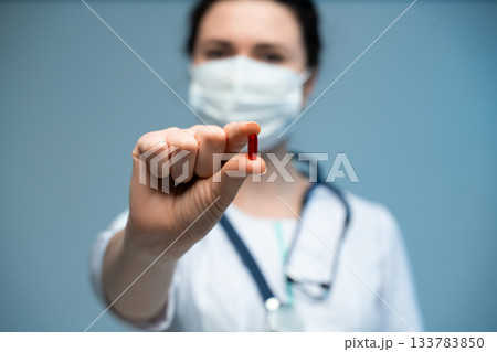 Female doctor in mask showing red capsule close to camera while wearing white uniform on blue background 133783850