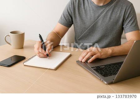 The man is working on a laptop and writing notes. The man is working on a laptop and writing notes. 133783970