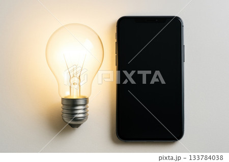 The light bulb glows next to the smartphone on a light background. The light bulb glows next to the smartphone on a light background. 133784038