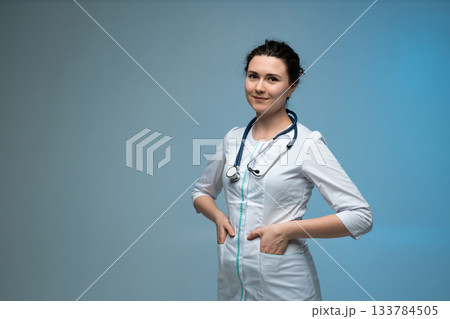 Confident female doctor with stethoscope posing with hands in pockets on blue background in uniform 133784505