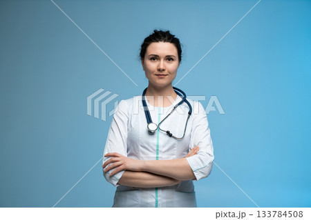 Confident female doctor with stethoscope standing with folded arms on blue background in medical uniform Confident female doctor with stethoscope standing with folded arms on blue background in medical uniform 133784508