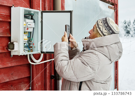 Meter reading shows resident woman with light hair using smartphone opposite snowy wall capturing meter digits in winter conditions. Meter reading shows resident woman with light hair using smartphone opposite snowy wall capturing meter digits in winter conditions. 133786743