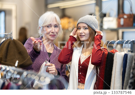 Young woman is fitting on and trying on warm hat in store, consulting with senior female assistant. 133787546