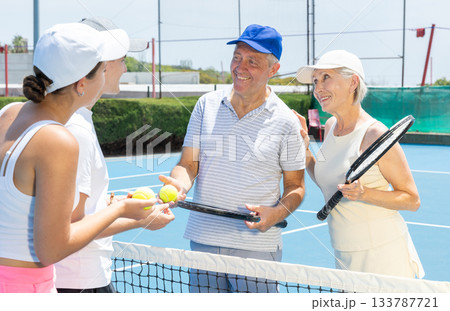Group of tennis players standing on open court on warm day with rackets and balls in hands, friendly chatting after match 133787721