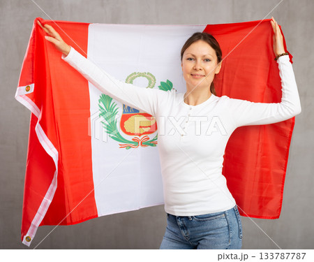 Smiling relaxed woman waving national flag of Peru while looking at camera at gray wall background 133787787