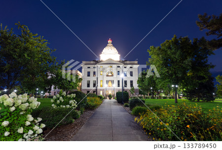 Illuminated South Dakota State Capitol building in Pierre at dusk. Walkway with flowers leads to the building, framed by trees under a clear blue sky Illuminated South Dakota State Capitol building in Pierre at dusk. Walkway with flowers leads to the building, framed by trees under a clear blue sky 133789450