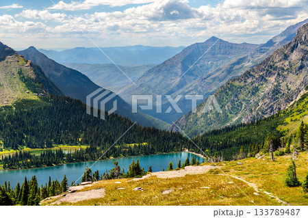 Hidden Lake sits in deep valley in Glacier National Park Montana. Alpine landscape features blue water and green pine forests surrounded by rugged mountain peaks Hidden Lake sits in deep valley in Glacier National Park Montana. Alpine landscape features blue water and green pine forests surrounded by rugged mountain peaks 133789487