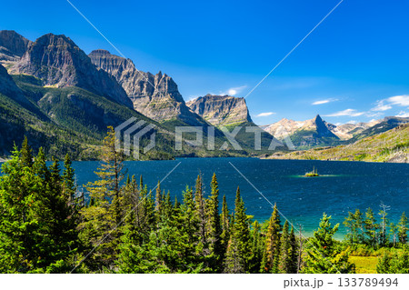 Panoramic view of the vibrant blue Saint Mary Lake, featuring Wild Goose Island, in Glacier National Park. The Rocky Mountains rise grandly under a clear blue sky, framed by pine trees 133789494