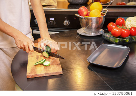 Person cutting fresh cucumbers on a wooden board surrounded by vegetables and fruits on the kitchen table. Healthy salad preparation and clean eating lifestyle at home. Person cutting fresh cucumbers on a wooden board surrounded by vegetables and fruits on the kitchen table. Healthy salad preparation and clean eating lifestyle at home. 133790061