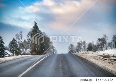 Snowy Road Scene With Wet Asphalt, Winter Trees, And Open Horizon Under Cloudy Sky 133790312