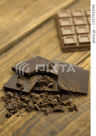 Dark chocolate squares with broken shards lying on wooden tabletop, showing segmented chocolate bar 133791049