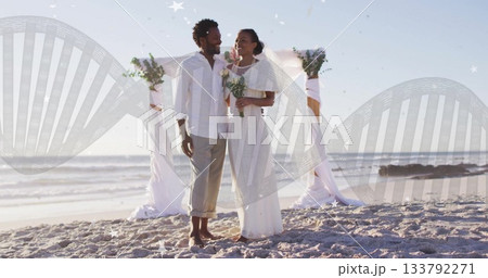 Standing bride and groom in wedding attire holding bouquets on sandy beach with floral arch Standing bride and groom in wedding attire holding bouquets on sandy beach with floral arch 133792271