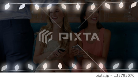 Sitting friends wearing gold, coral dresses on teal bench in lounge, with smartphones and lights 133793070