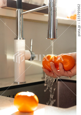 Person Washes Two Mandarin Oranges Under Running Water in Kitchen Sink 133795662