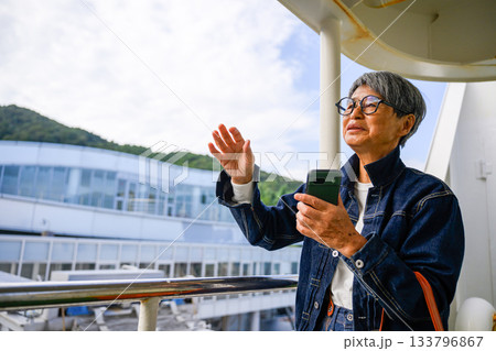 スマートフォンを持ち、出港するフェリーのデッキで手を振るシニア女性 スマートフォンを持ち、出港するフェリーのデッキで手を振るシニア女性 133796867