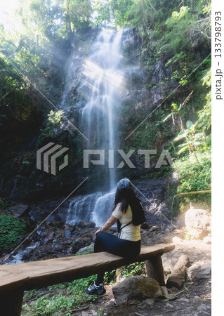 Serene Woman Enjoys Nature by Majestic Waterfall Surrounded by Lush Greenery in Tranquil Forest Setting 133798793