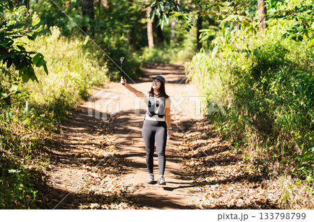 Young woman enjoys outdoor photography on hiking trail in forest, capturing moments with selfie stick during sunny day surrounded by trees and nature Young woman enjoys outdoor photography on hiking trail in forest, capturing moments with selfie stick during sunny day surrounded by trees and nature 133798799