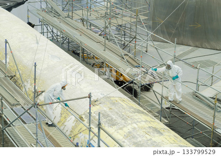 Workers prepare a large aircraft section for painting at a maintenance facility while using scaffolding and protective gear in a well-lit environment Workers prepare a large aircraft section for painting at a maintenance facility while using scaffolding and protective gear in a well-lit environment 133799529