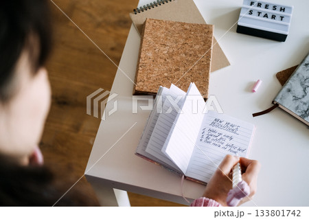 Person writes New Year resolutions list in small notebook on white table next to FRESH START light box. New Year resolutions, micro-habits, daily rituals, consistency. 133801742