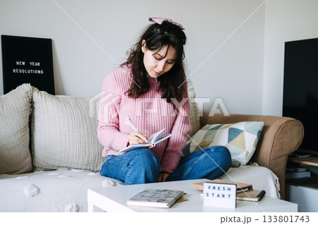 Young adult woman sits on sofa writing goals in small notebook next to letter board displaying NEW YEAR RESOLUTIONS. New Year resolutions, Micro-goals, goal setting, self improvement. 133801743