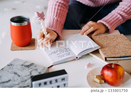 Person writes New Year resolutions list in small notebook using pearl pen next to coffee tumbler and apple. New Year resolutions, goal setting, journaling, healthy lifestyle. Person writes New Year resolutions list in small notebook using pearl pen next to coffee tumbler and apple. New Year resolutions, goal setting, journaling, healthy lifestyle. 133801750