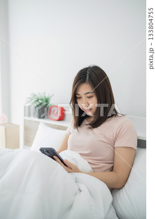 Young woman relaxing in bed using smartphone while wrapped in blanket in cozy bedroom setting with decor, suitable for lifestyle themes and digital communication concepts Young woman relaxing in bed using smartphone while wrapped in blanket in cozy bedroom setting with decor, suitable for lifestyle themes and digital communication concepts 133804755
