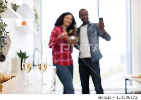 A couple is in their kitchen, with the man taking a selfie while the woman holds a bowl. Two glasses of wine are on the counter. A couple is in their kitchen, with the man taking a selfie while the woman holds a bowl. Two glasses of wine are on the counter. 133806172