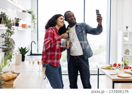 A diverse couple smiles while taking a selfie in their modern kitchen, enjoying a salad and wine. 133806176