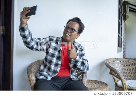 Indonesian southeast asian man takes a selfie using a cellphone while sitting on a chair outdoors Indonesian southeast asian man takes a selfie using a cellphone while sitting on a chair outdoors 133806707