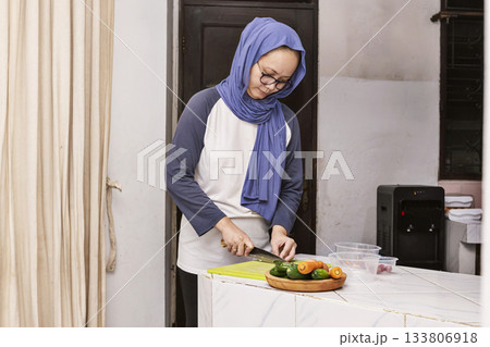 Indonesian woman wearing a blue scarf is cutting vegetables on a cutting board 133806918