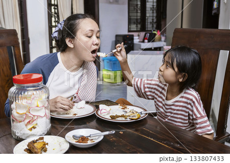 Asian woman and a child are eating in dinning table. Daughter feeding her mother while eating together. Asian woman and a child are eating in dinning table. Daughter feeding her mother while eating together. 133807433