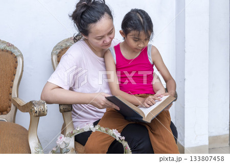Woman and a child are sitting on a chair reading a book 133807458