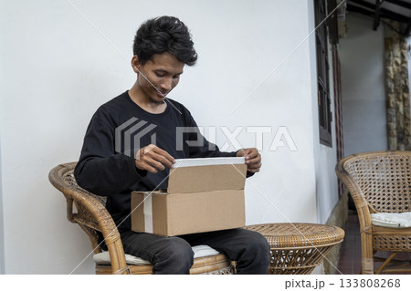 Indonesian southeast asian man on a chair opening a cardboard box and looking inside. Delivery service Indonesian southeast asian man on a chair opening a cardboard box and looking inside. Delivery service 133808268