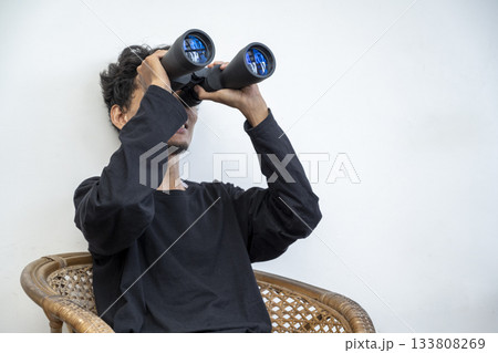 Indonesian southeast asian man sitting on a chair. Using a new pair of binoculars from the package. Delivery service Indonesian southeast asian man sitting on a chair. Using a new pair of binoculars from the package. Delivery service 133808269