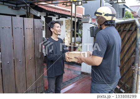 Indonesian southeast asian man receives a cardboard package from a delivery person wearing a helmet. Delivery service 133808299